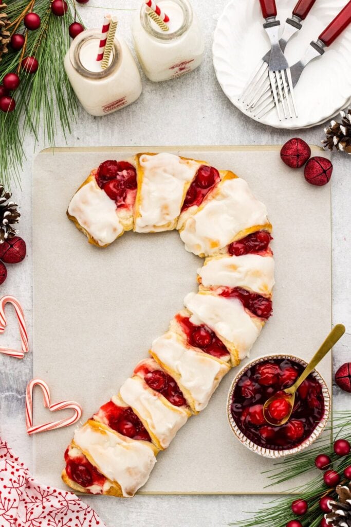 Candy cane-shaped pastry with cherry filling and icing on parchment, surrounded by pine branches, ornaments, milk bottles with striped straws, and festive tableware.