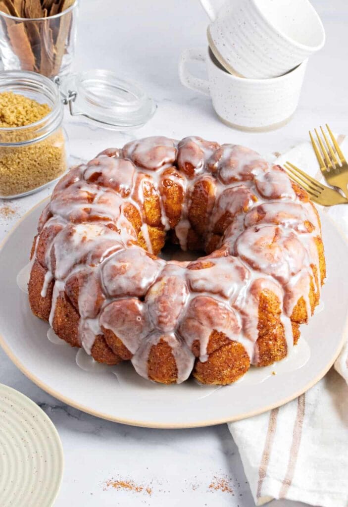 Glazed monkey bread of cinnamon-sugar dough pieces on white plate, surrounded by brown sugar jar, cinnamon sticks, stacked cups, gold forks, and striped napkin on marble surface.