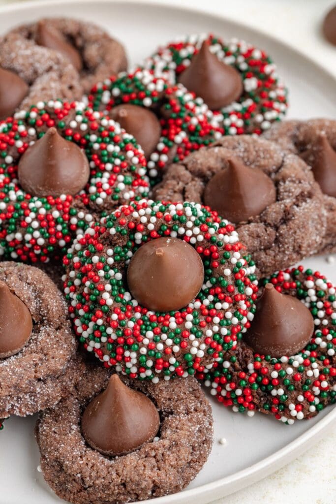 Chocolate cookies topped with Hershey’s Kiss, decorated with red, white, and green sprinkles or sugar, arranged on light plate.