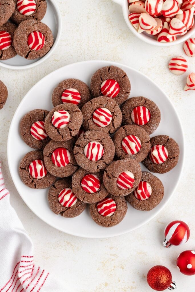 Chocolate cookies topped with red-and-white peppermint candies, arranged on plate with bowls of extra cookies and candies, festive cloth, ornaments, and holiday accents nearby.