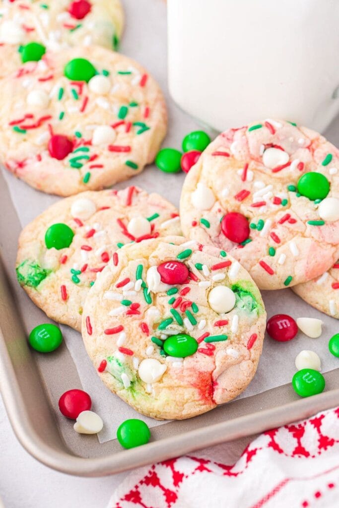 Tray of golden cookies with red-green sprinkles, white chips, and M&M candies on parchment, with milk glass and patterned cloth nearby.