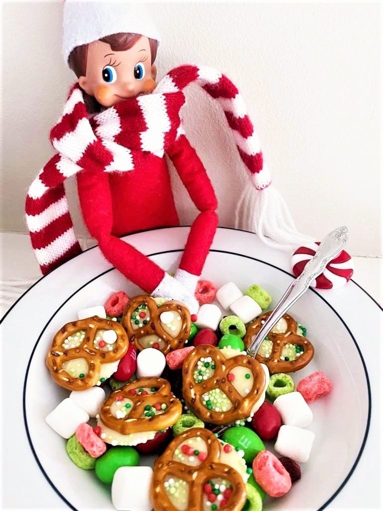 Elf on the Shelf doll with red outfit and striped scarf behind bowl of pretzels topped with white chocolate and sprinkles, surrounded by marshmallows, candies, and cereal.