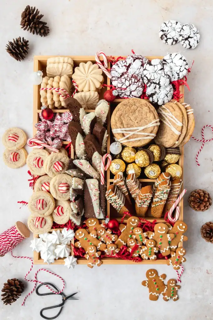 Wooden tray with assorted holiday cookies—gingerbread men, peppermint-topped sugar cookies, biscotti, crinkle cookies, shortbread, iced rounds—plus truffles, meringues, and candy canes, surrounded by pinecones, twine, ornaments, and scissors.