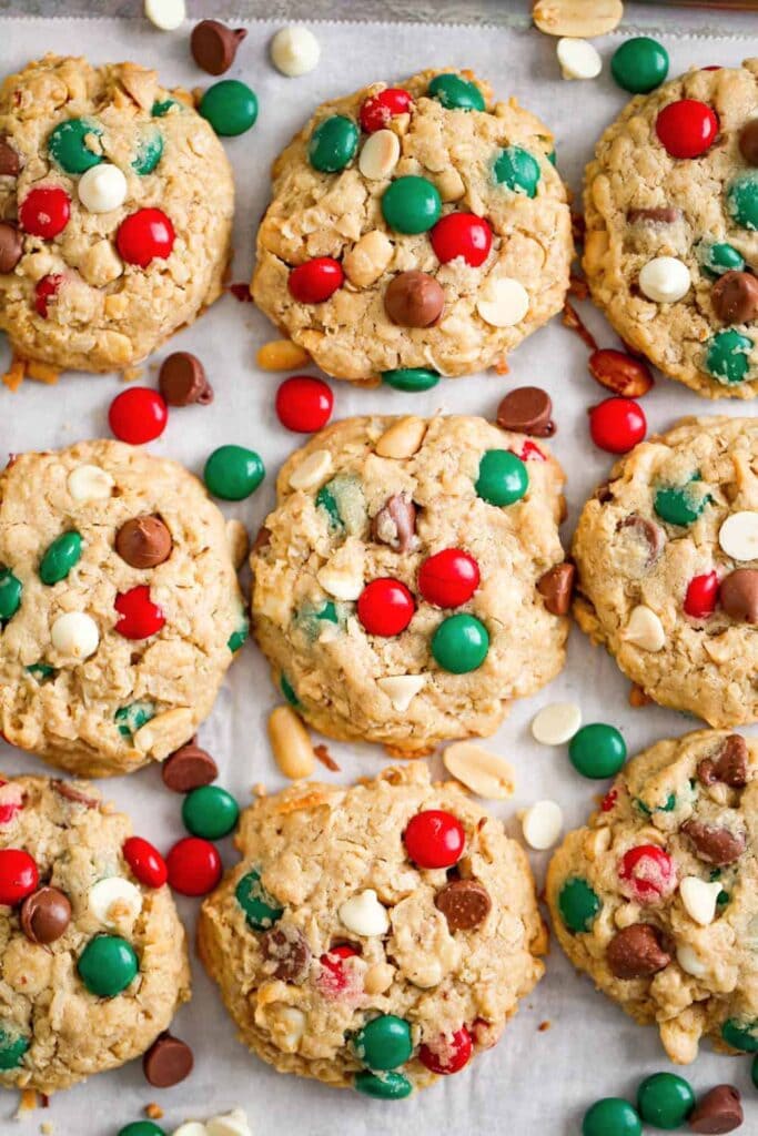 Chunky golden cookies with red-green candies, chocolate chips, and peanuts on parchment-lined baking sheet.