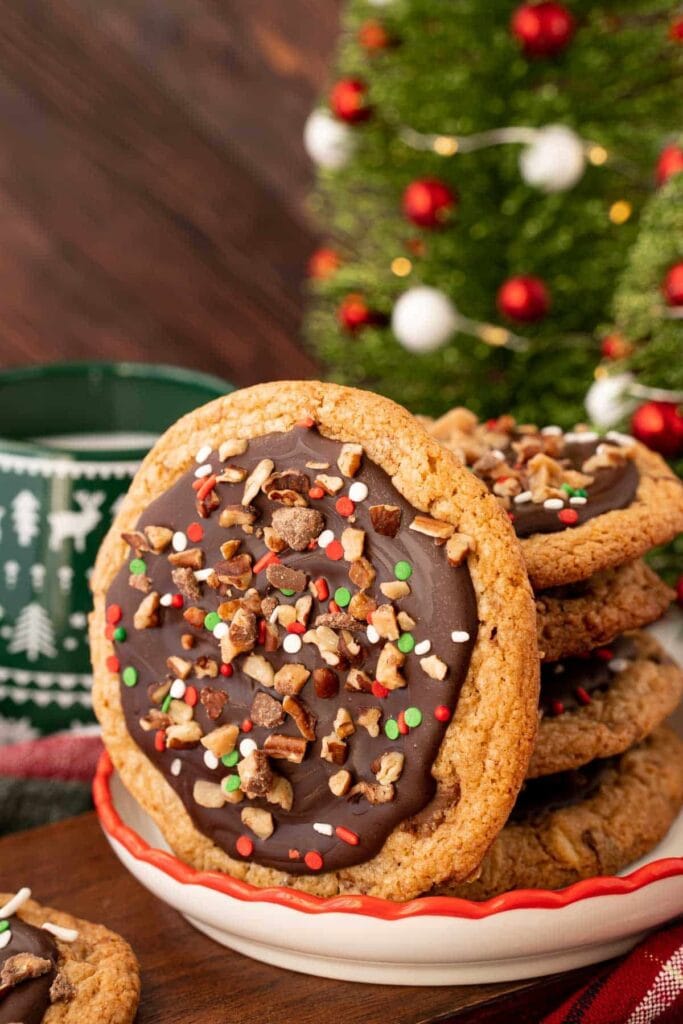 Stack of large cookies topped with chocolate frosting, nuts, and red, green, white sprinkles on white plate with red rim, festive mug and Christmas tree in background.