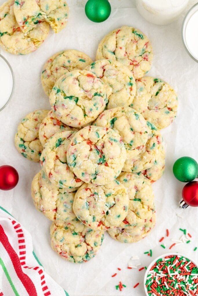 Golden cookies with red-green sprinkles on white surface, surrounded by ornaments, bowls of milk and sprinkles, and striped holiday cloth.