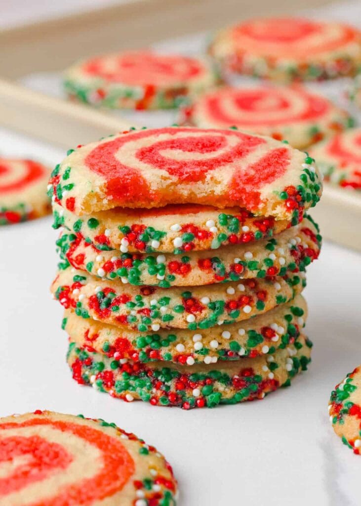 Stack of sugar cookies with red swirl tops and edges coated in red, green, and white sprinkles, one with bite taken, with more cookies on tray in background.