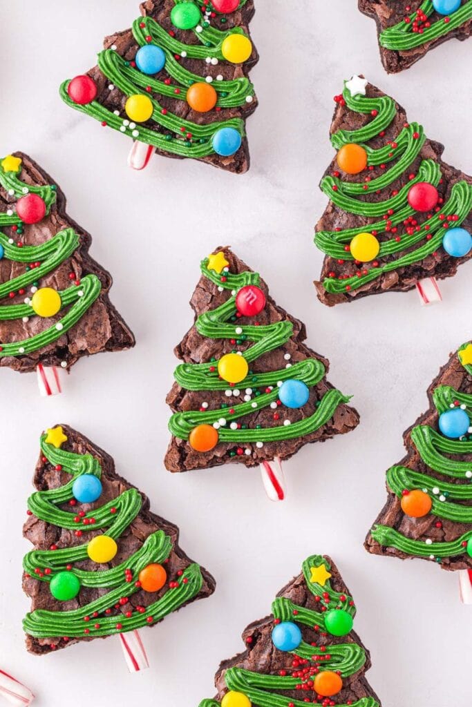 Brownies cut into Christmas tree shapes, decorated with green frosting garlands, colorful candy ornaments, and candy cane trunks, arranged on white surface.