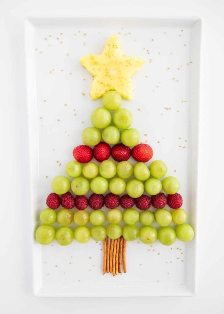 Christmas tree-shaped fruit arrangement with grape rows, strawberry and raspberry garland, pineapple star topper, and pretzel stick trunk on white plate.