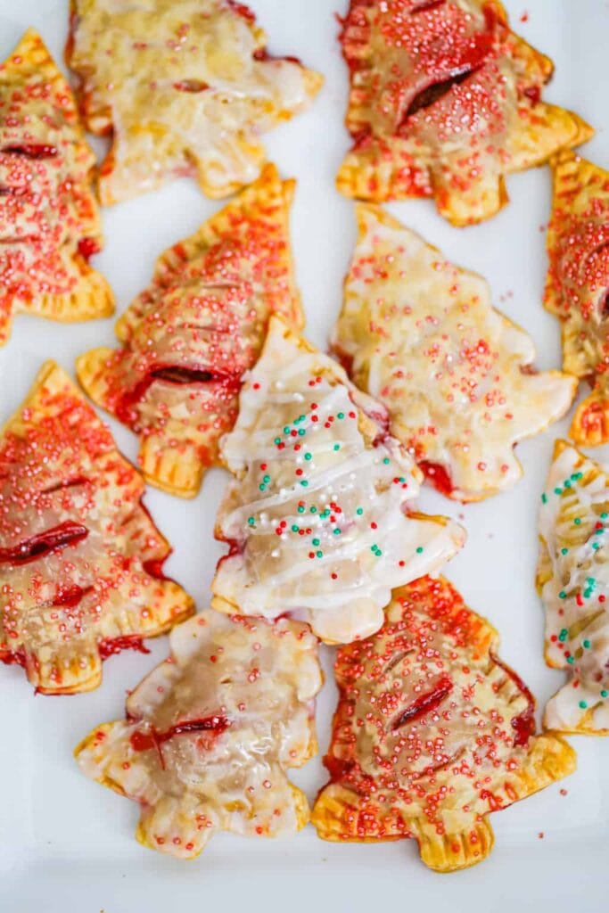 Golden-brown Christmas tree pastries filled with red fruit, decorated with icing and red, white, green sprinkles, arranged on white surface.