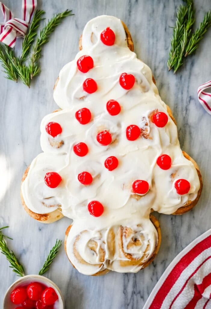 Cinnamon rolls arranged as Christmas tree with white icing and red cherries, decorated with rosemary sprigs and striped ribbons on marble surface.