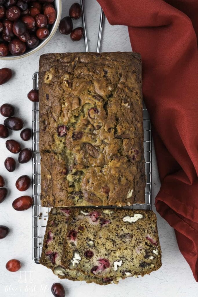 Loaf of cranberry nut bread with golden crust, sliced to show cranberries and nuts inside, on cooling rack with scattered cranberries, bowl of berries, and red cloth nearby.