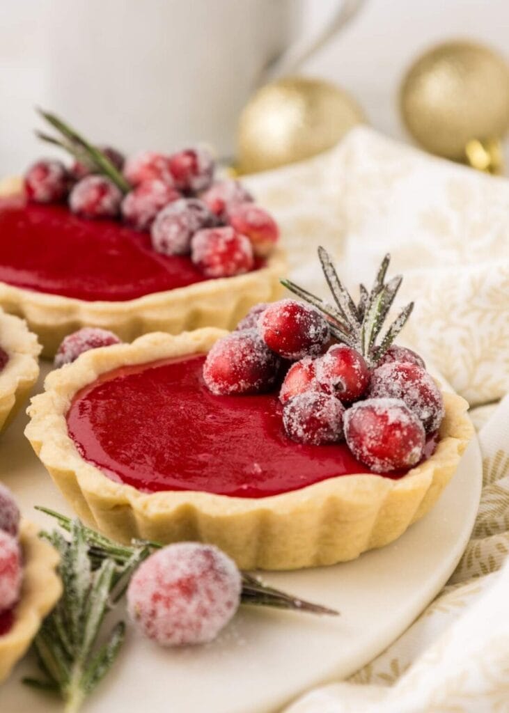 Small tarts with glossy red filling, topped with sugared cranberries and rosemary sprigs, set on light surface with patterned cloth and gold ornaments.
