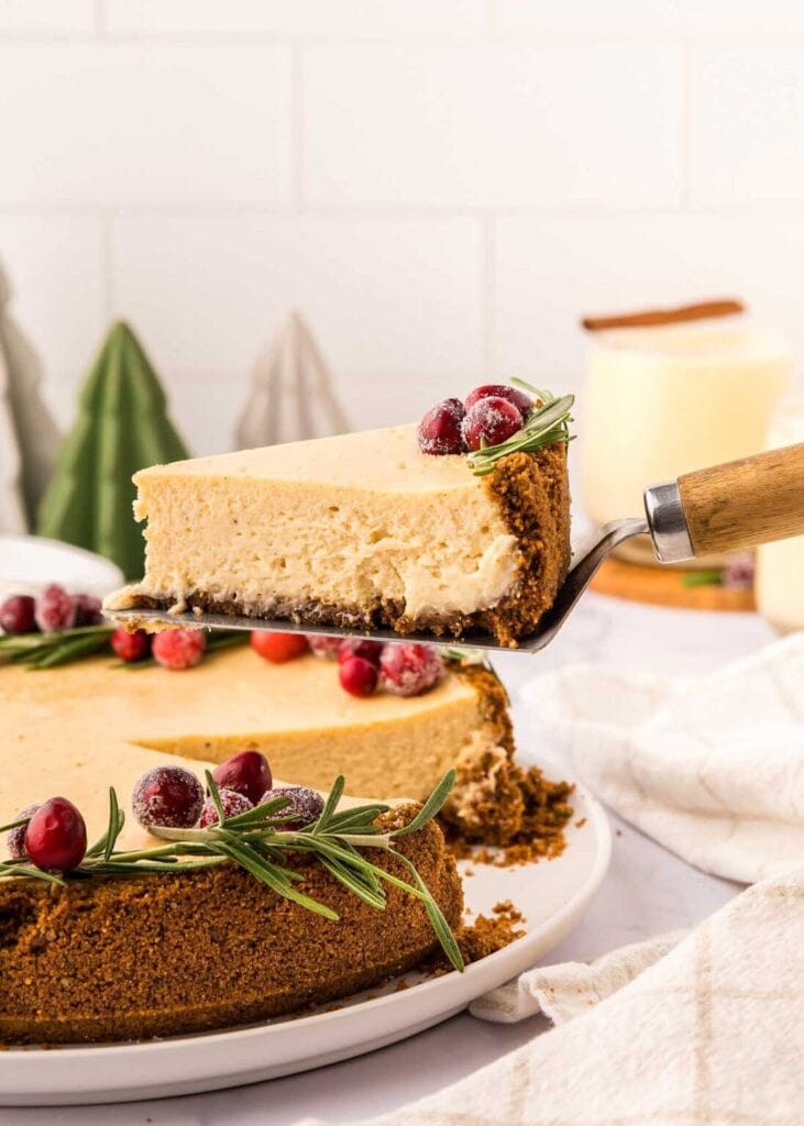Cheesecake slice with sugared cranberries and rosemary garnish lifted from whole cake with graham crust, decorated similarly, on cozy holiday table.