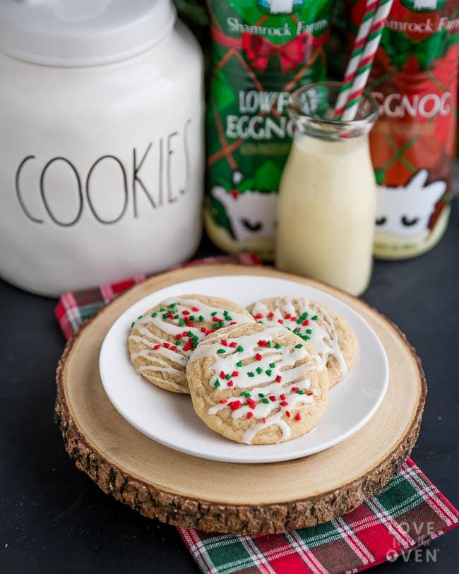 Plate of iced cookies with red-green sprinkles on wooden board, beside cookie jar, eggnog cartons, and glass bottle with striped straw on plaid cloth.