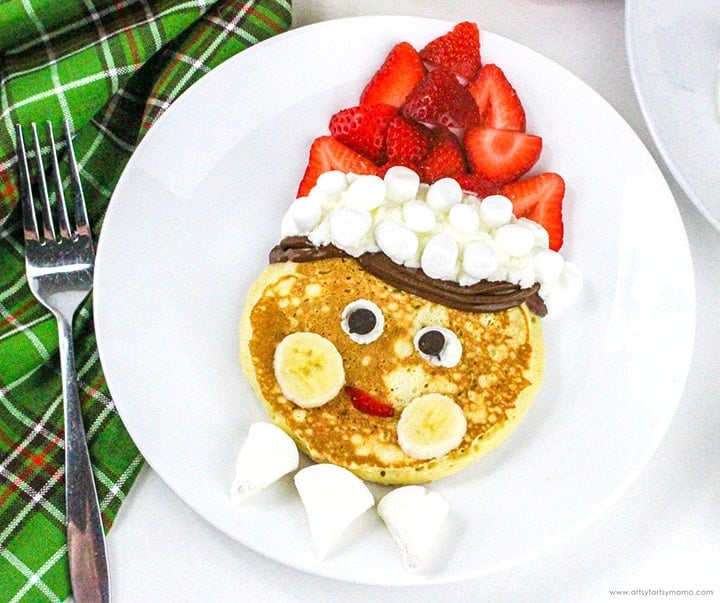 Pancake decorated as playful face with banana cheeks, chocolate hair, marshmallow and strawberry hat, candy eyes, and red mouth on white plate with plaid cloth and fork.