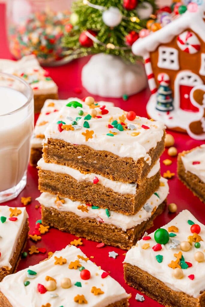 Stacked frosted dessert bars with colorful sprinkles on red surface, beside glass of milk, small Christmas tree, and gingerbread house ornament.