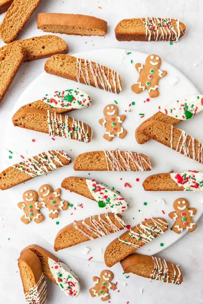 Gingerbread biscotti decorated with icing and sprinkles, some dipped with candy cane and tree toppings, arranged with smiling gingerbread cookies on white surface.