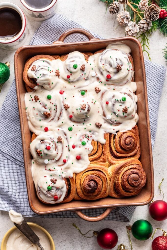 Baking dish of cinnamon rolls, top glazed with icing and festive sprinkles, bottom showing golden spirals, surrounded by coffee cups, icing bowl, cloth, pinecones, greenery, and ornaments.