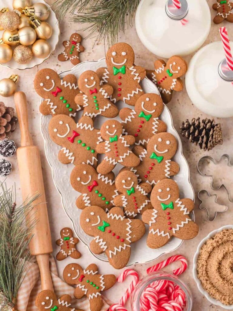 Plate of gingerbread people cookies with icing and candy buttons, surrounded by candy canes, pinecones, rolling pin, cutters, sugar bowl, milk bottles with striped straws, ornaments, and evergreen branches.