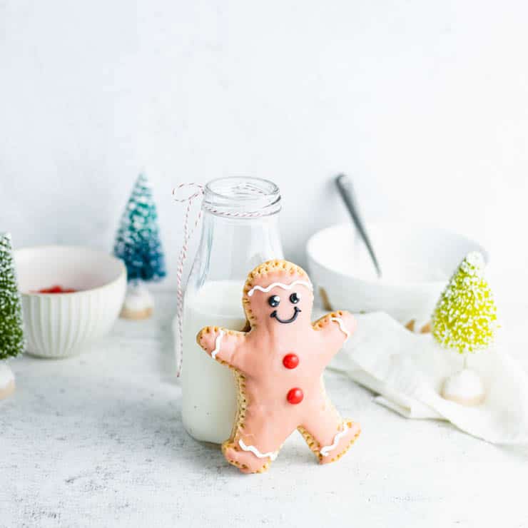 Gingerbread man with pink icing and red candy buttons leaning against milk bottle tied with red-white string, surrounded by festive trees and bowls of candies.