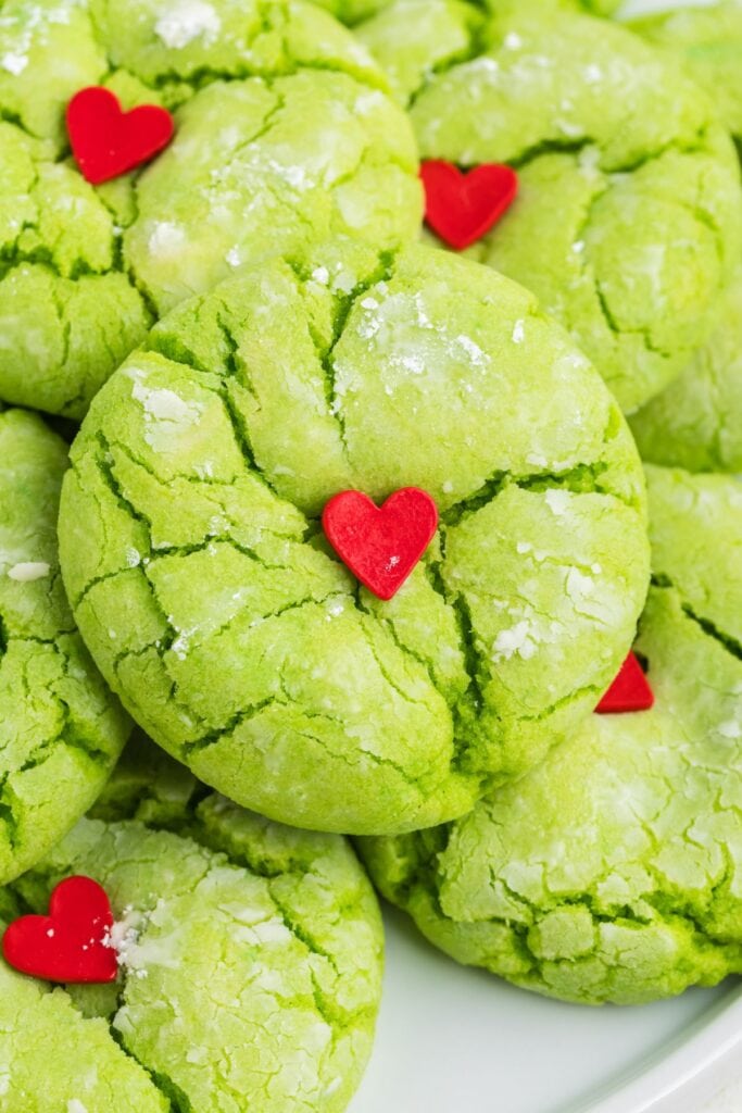 Bright green cracked cookies dusted with powdered sugar, each topped with small red heart decoration, arranged on white plate.