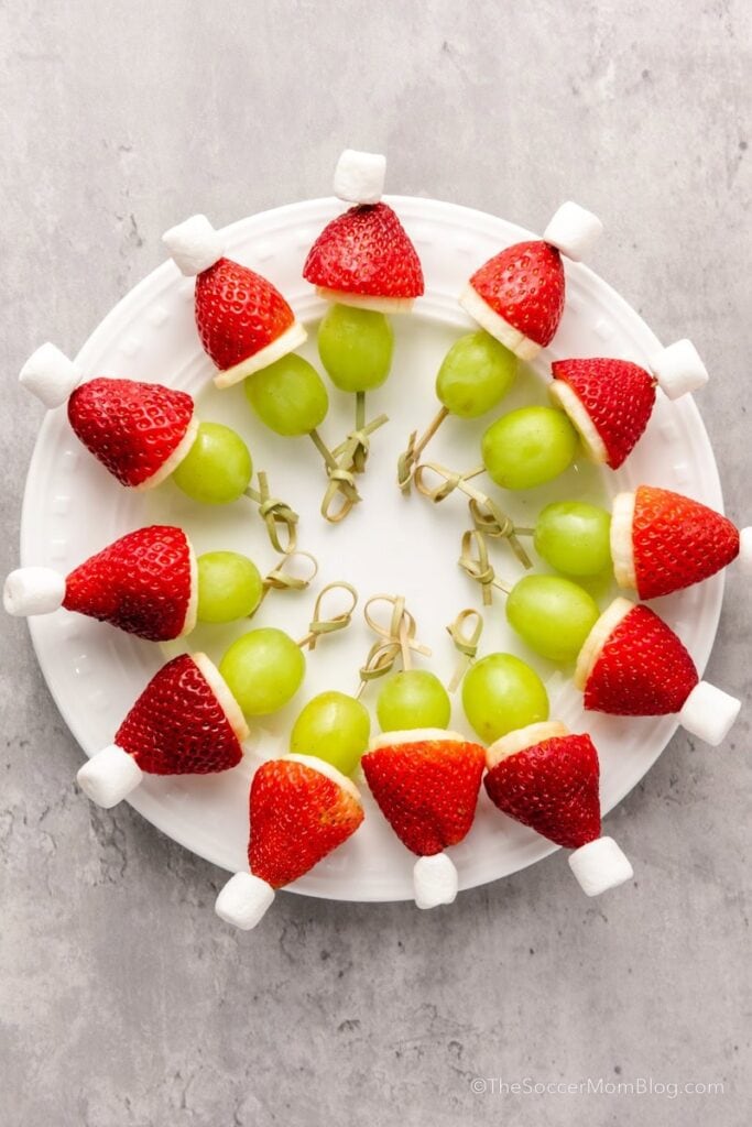 Fruit skewers with grape, banana, strawberry, and marshmallow arranged in circle on white plate, resembling Santa hats.