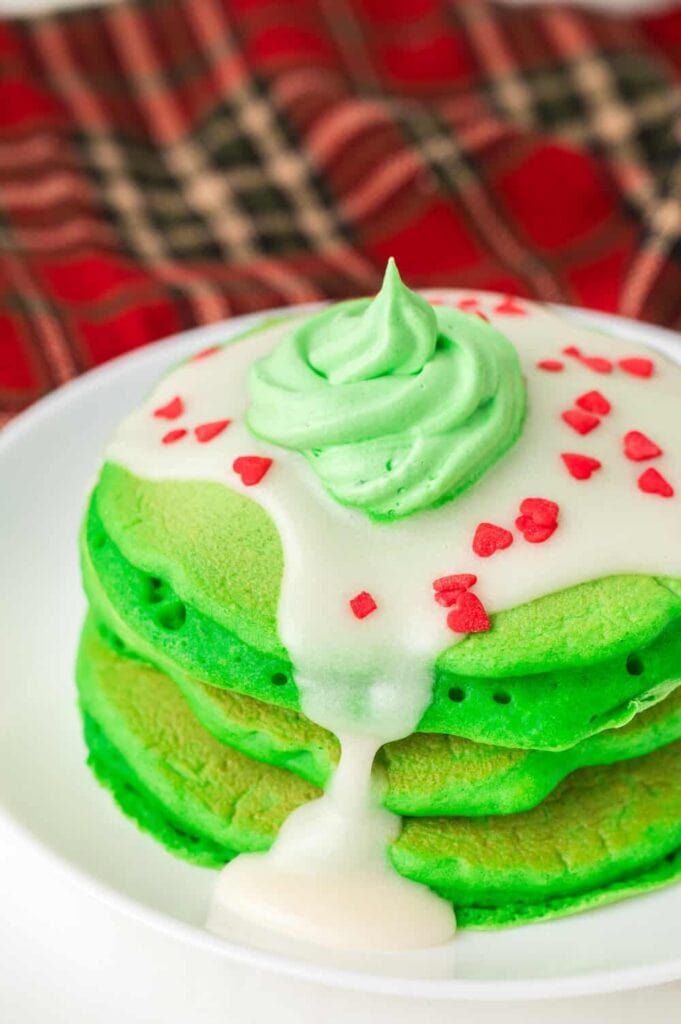 Stack of green pancakes with frosting swirl, white icing drizzle, and red heart sprinkles on white plate with plaid fabric background.