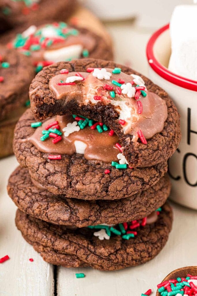 Stack of chocolate cookies with red-green sprinkles, marshmallow filling, and melted chocolate topping beside white mug with marshmallows.