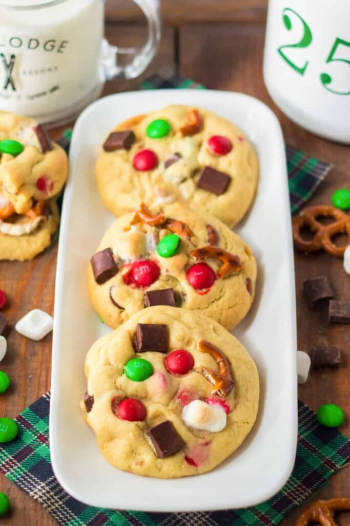 Large cookies with red-green candies, marshmallows, pretzels, and chocolate chunks on white plate with plaid cloth, surrounded by holiday treats and mugs of milk.