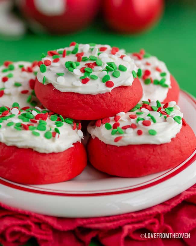 Stack of red cookies with white frosting and red-green sprinkles on white plate with red rim, set on red cloth with green holiday background.