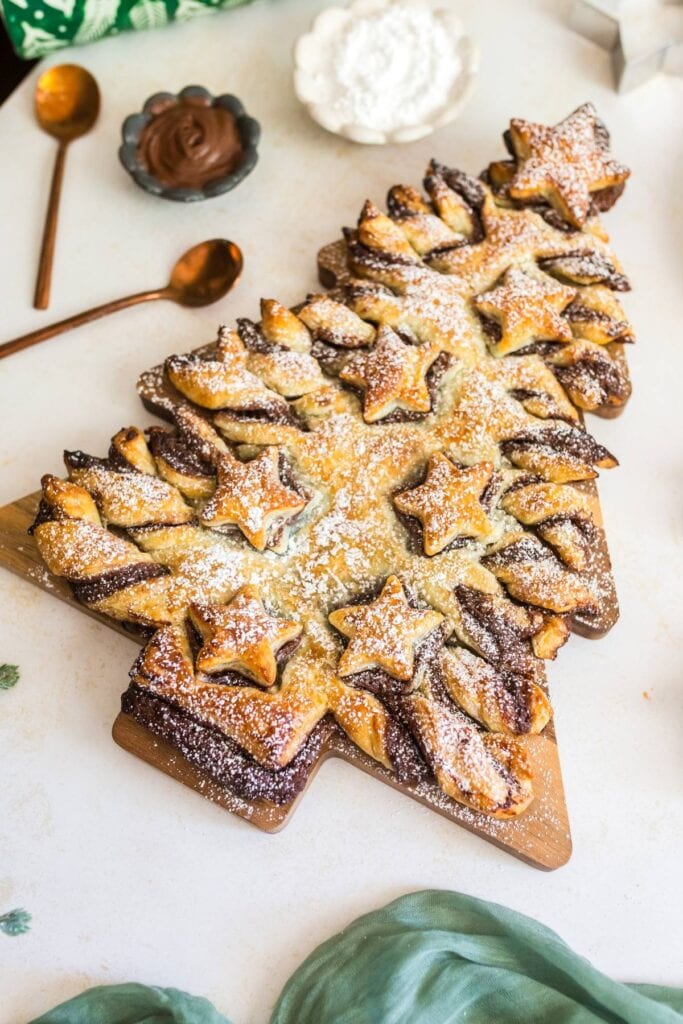 Christmas tree-shaped pastry with chocolate filling, dusted with powdered sugar, decorated with star-shaped dough, on wooden board with spoons, chocolate spread, and powdered sugar bowl.