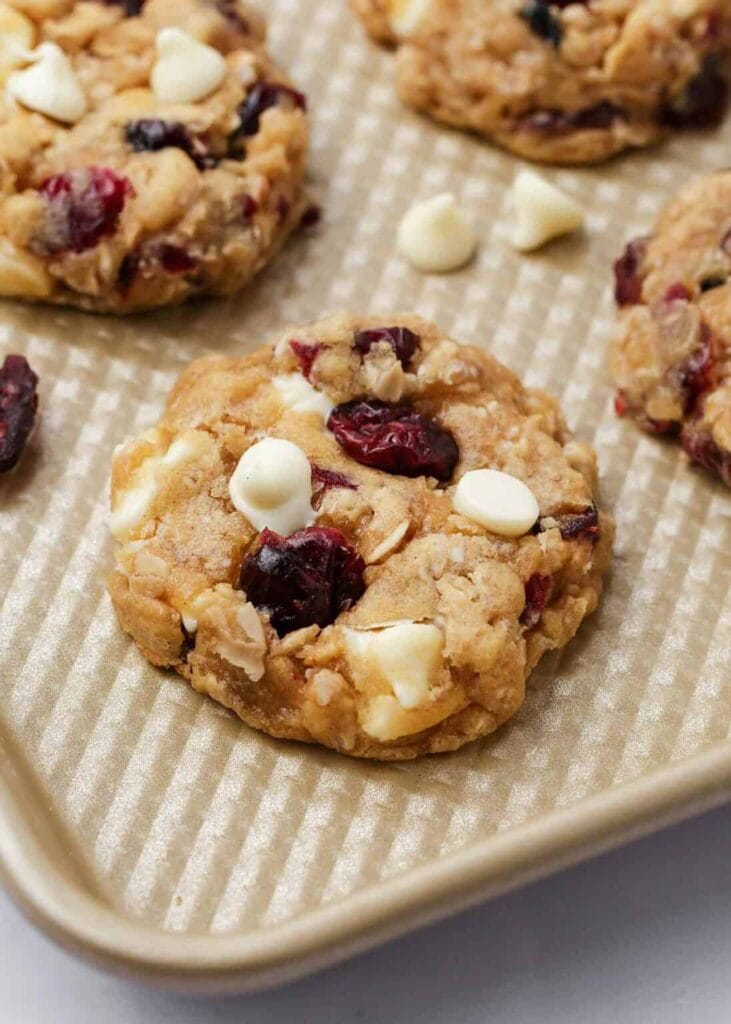 Golden oatmeal cookies with cranberries and white chocolate chips on textured baking sheet.