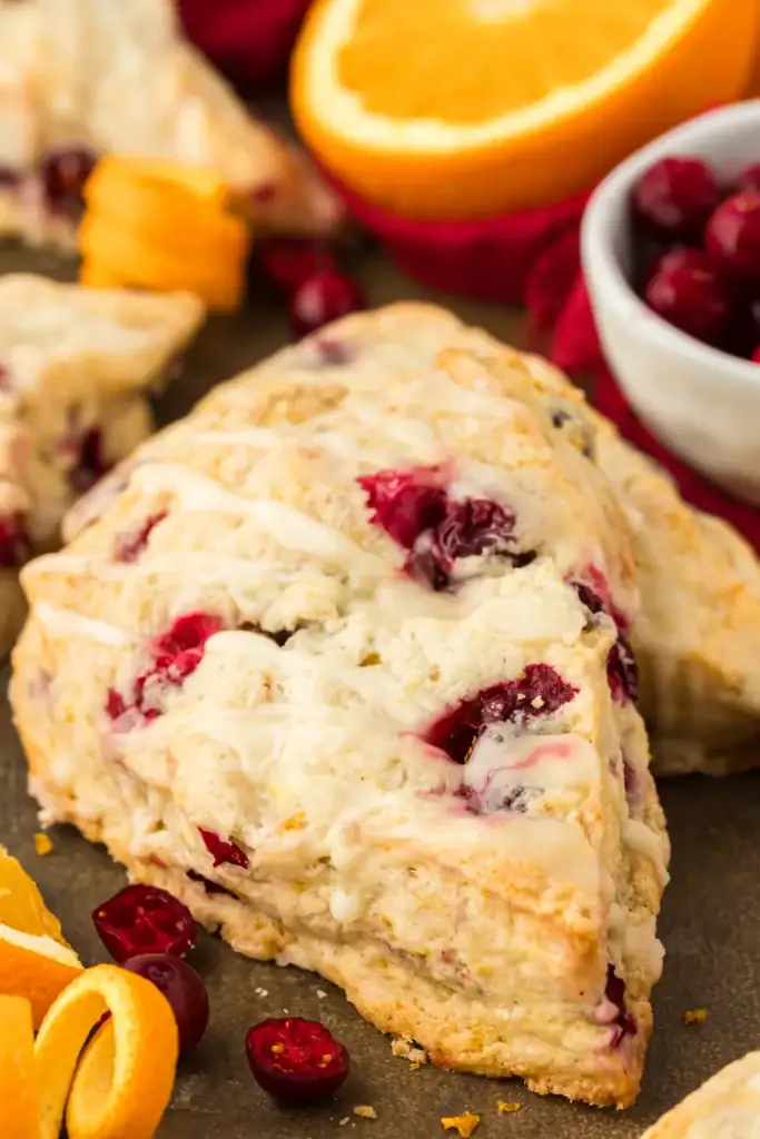 Golden cranberry-orange scone with icing drizzle, surrounded by cranberries and orange slices on white surface.