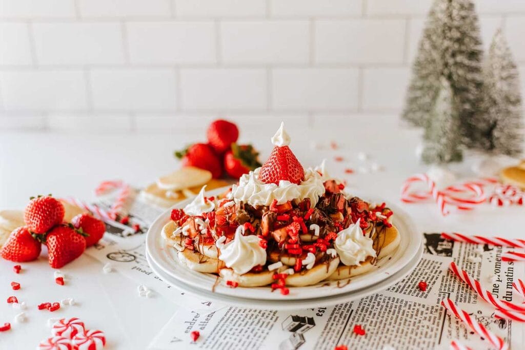 Waffles topped with whipped cream, strawberries, chocolate drizzle, and peppermint candies, surrounded by candy canes, fresh strawberries, and small Christmas trees.