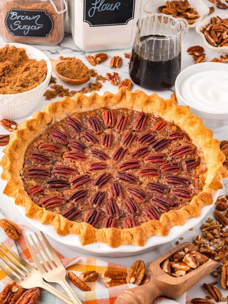 Golden pecan pie with circular nut topping on checkered cloth, surrounded by baking ingredients—brown sugar, flour, syrup, white sugar, chopped pecans—and gold forks.