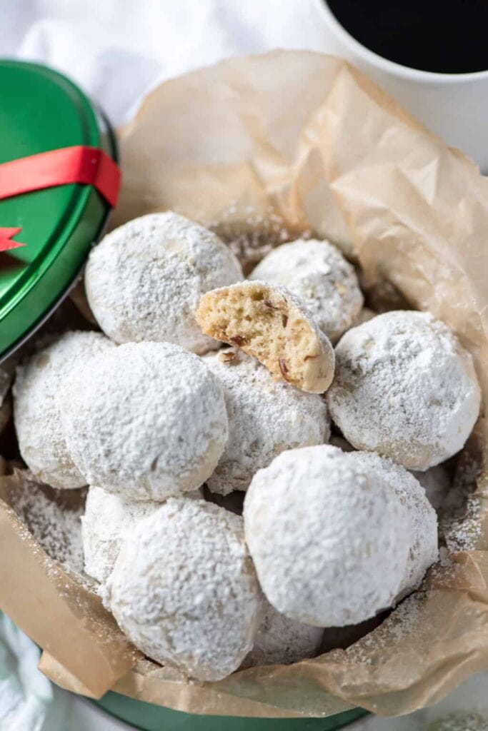 Powdered sugar-coated snowball cookies in parchment-lined tin, one bitten to reveal nutty interior, with green tin ribbon and coffee cup nearby.