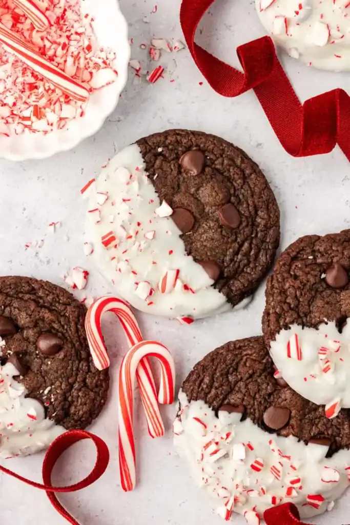 Chocolate cookies dipped in white chocolate, sprinkled with crushed peppermint, arranged with candy canes, red ribbon, and bowl of peppermint pieces.