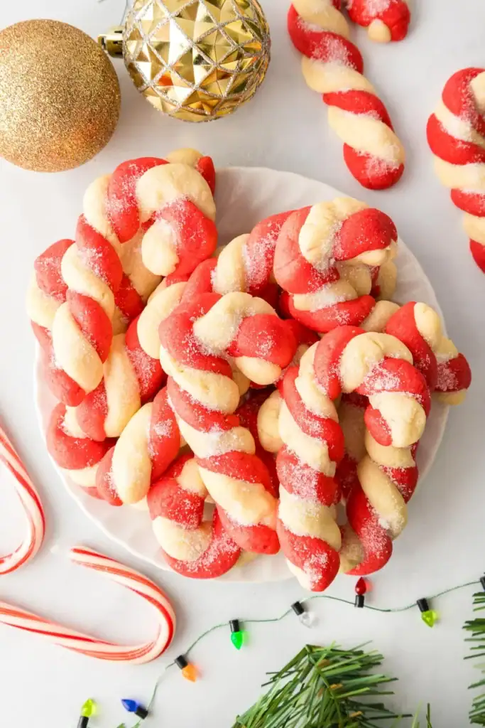 Plate of red-and-white twisted candy cane cookies dusted with sugar, surrounded by ornaments, real candy cane, string lights, and pine branches.