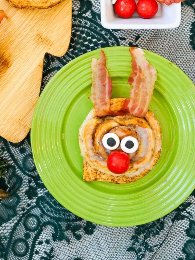 Cinnamon roll with icing styled as reindeer face, bacon antlers, candy eyes, and red nose on green plate, with cherry tomatoes and cutting board in background.