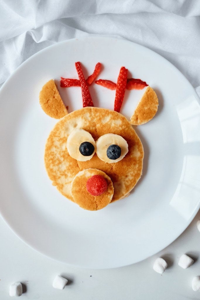 Pancake reindeer face with banana-blueberry eyes, raspberry nose, strawberry antlers, arranged on white plate with marshmallows scattered around.