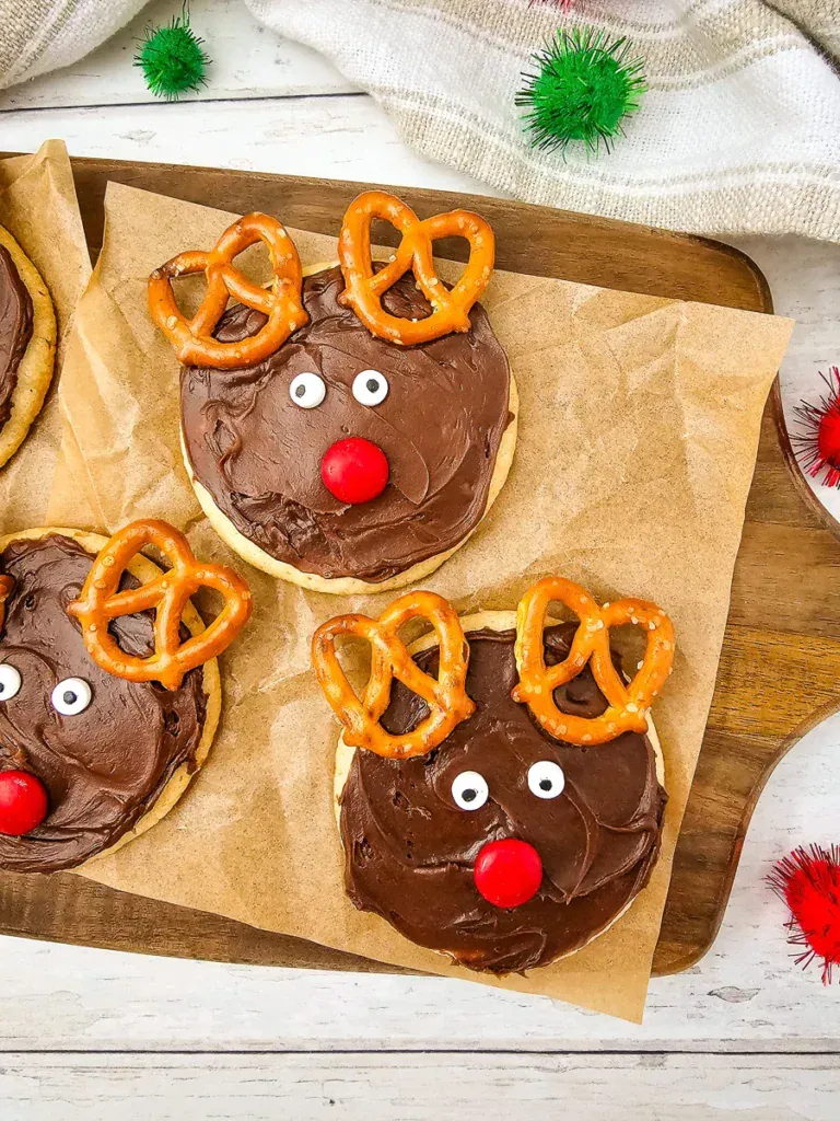 Round cookies decorated as reindeer with chocolate icing, pretzel antlers, candy eyes, and red noses on parchment paper with festive pom-poms.