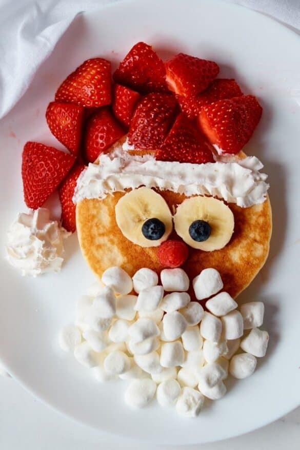 Round pancake styled as Santa face with banana-blueberry eyes, berry nose, whipped cream trim, strawberry hat, and marshmallow beard on white plate.