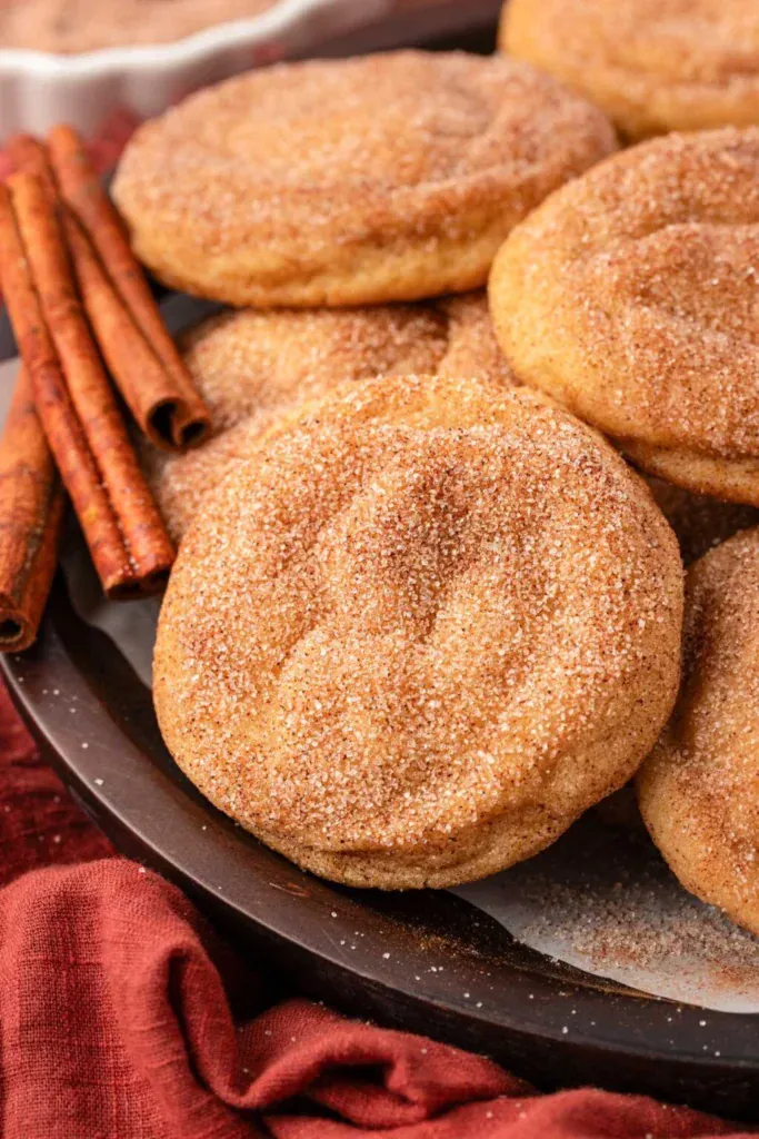 Golden snickerdoodle cookies coated in cinnamon sugar on dark plate with cinnamon sticks and red cloth accent.
