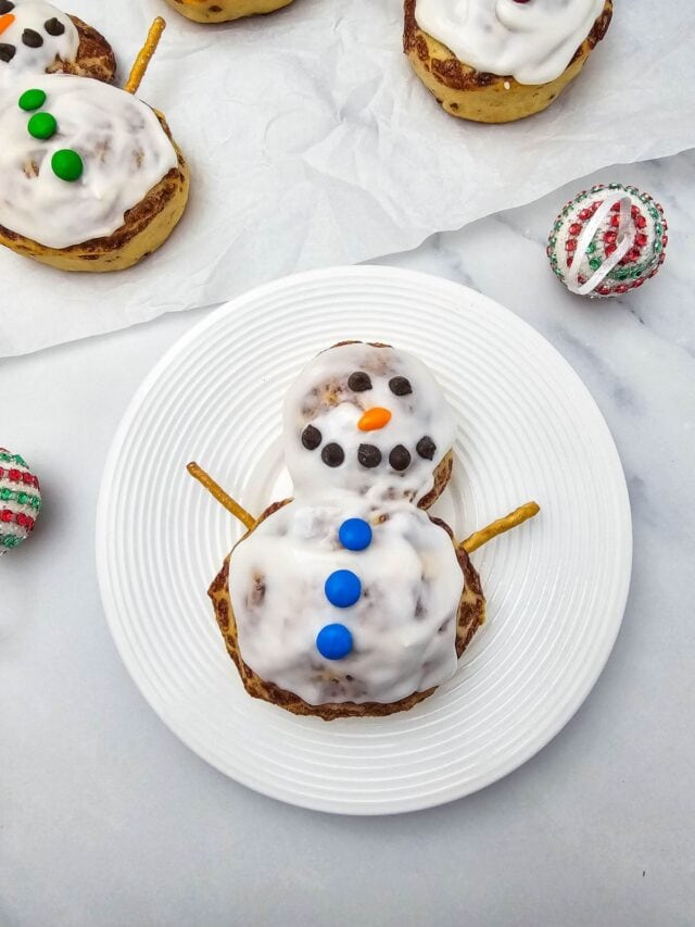 Two stacked iced cinnamon rolls shaped as snowman, decorated with chocolate chip face, orange candy nose, blue buttons, and pretzel arms, with more rolls and ornaments in background.
