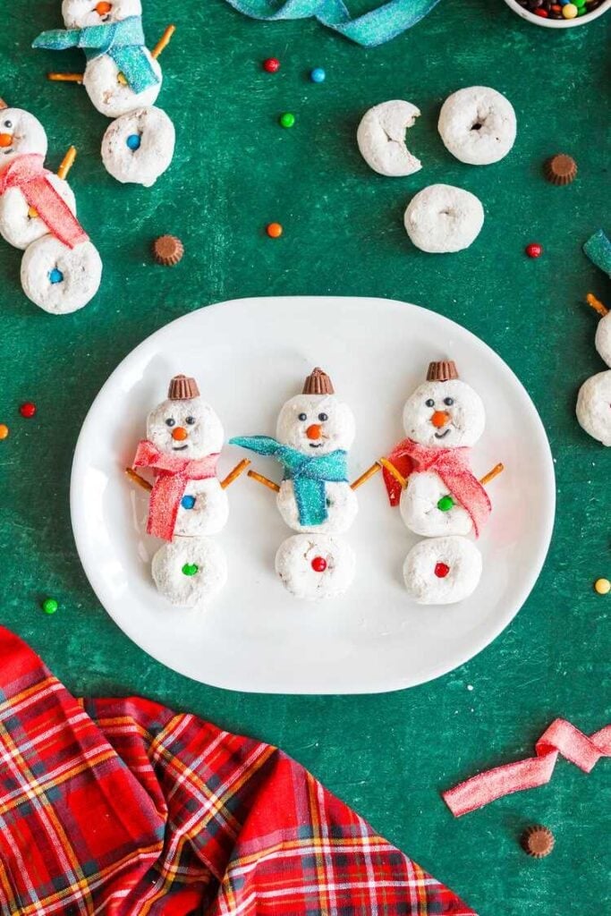 Three stacked powdered donut snowmen with candy faces, gummy scarves, peanut butter cup hats, and pretzel arms on white plate, surrounded by festive candies and plaid cloth.