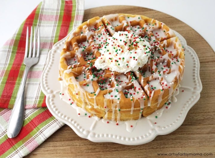 Stack of waffles with icing drizzle, whipped cream, and red-green sprinkles on white plate beside plaid napkin and fork.