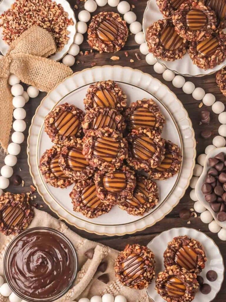 Thumbprint cookies with caramel centers, chocolate drizzle, and pecan edges, arranged on plates with bowls of chocolate, pecans, and rustic festive decorations.