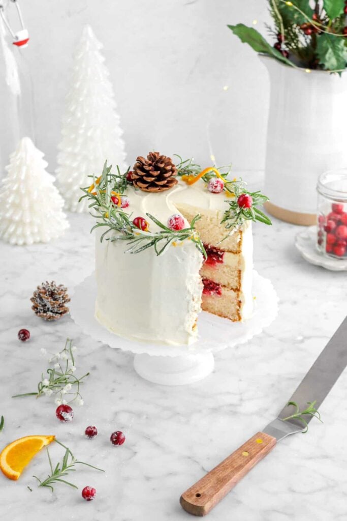 Three-layer vanilla cake with berry filling and white frosting, topped with pinecone, sugared cranberries, rosemary, and orange twist, on cake stand with holiday decorations.